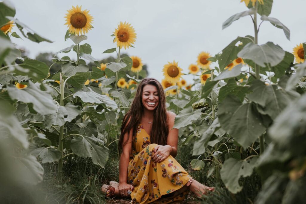 A woman with long brown hair and brown skin sits on the ground in a sunflower field. She wears a yellow flowered dress, crosses her legs, and holds onto them. She's laughing and her eyes are closed, so the focal point is her bright, wide smile. Endless sunflowers are in the background under a gray sky.