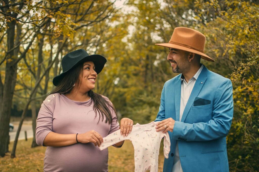 A man and a woman stand underneath tall green trees in early autumn. Slightly yellow leaves cover the nature trail behind them. The woman wears a black wide-rimmed hat and a purple shirt. The man wears an orange wide-rimmed hat, a white collared shirt, a light blue suit jacket and white pants. The woman is pregnant. They hold a baby's onesie between them and smile as they look at each other - how to prepare for labor