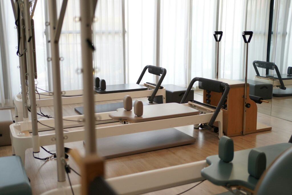 Pilates reformer machines with attached springs, straps, and padded platforms inside a bright studio with large windows and sheer curtains.