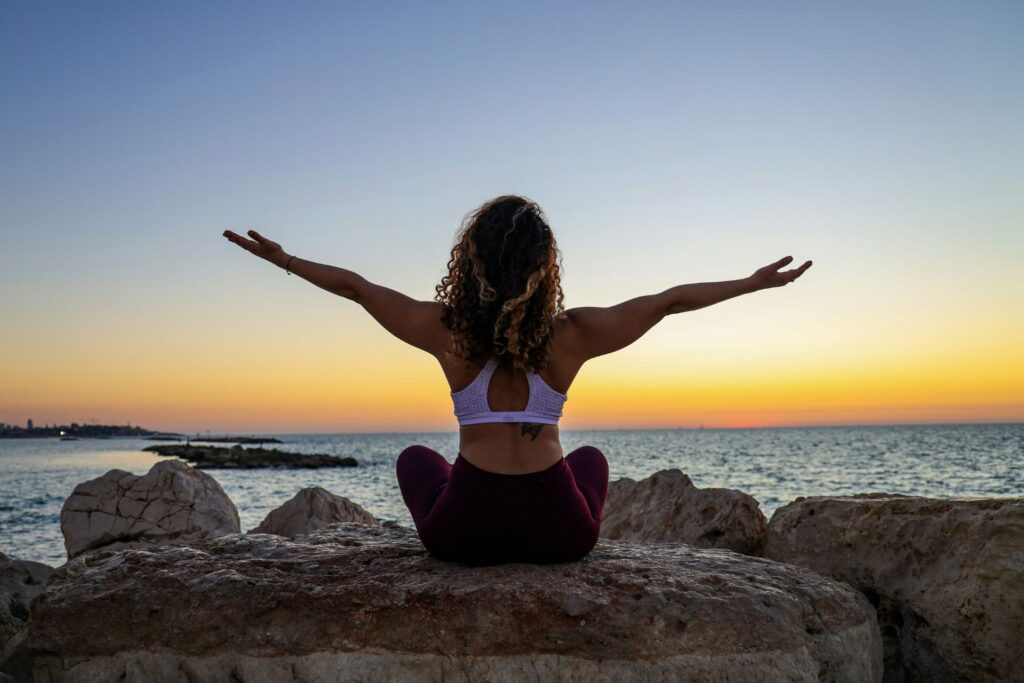 A woman sitting cross-legged on a large rock by the sea, facing away from the camera with arms outstretched, practicing yoga during sunset.