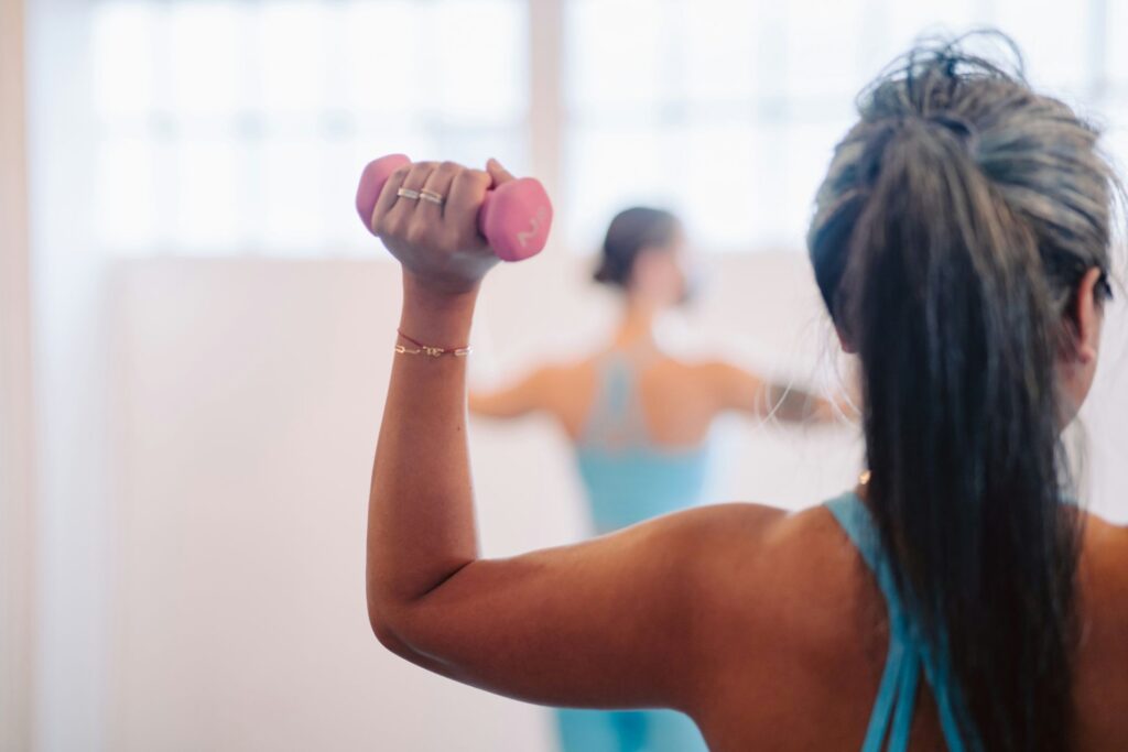 Woman in a teal tank top lifting a pink dumbbell, viewed from behind, with reflection in a mirror.