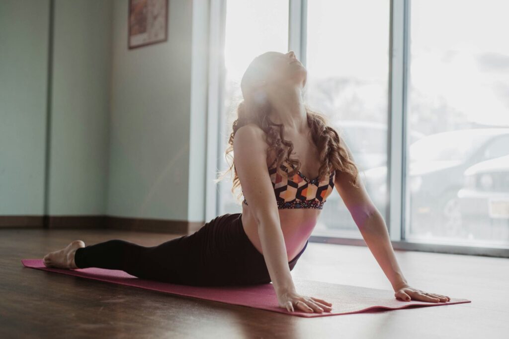 A woman with long curly hair wearing a colorful sports bra and black leggings practicing yoga on a pink mat in a room with wooden floors and large windows. She is doing the upward-facing dog pose with her head tilted back and eyes closed, sunlight streaming in from the window.