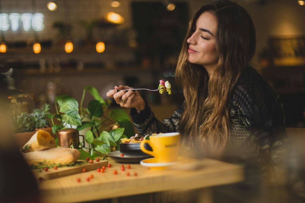 A woman sits in a restaurant in a black long-sleeved sweater. She holds a fork in the air with spiral pasta on it. Her eyes are closed and she's smiling. More pasta is in a bowl in front of her next to a yellow mug, a plant, red berries and a cutting board covered in bread. The rest of the background is blurry, but warm orange lights are noticeable.