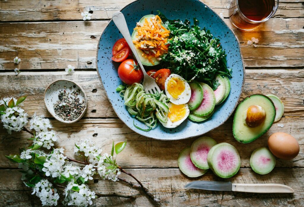 A wooden table is covered in dogwood flowers, various sliced fruits and a bowl of kale, spiralized zucchini, tomatoes, half an avocado, eggs, cheese and a fork. 