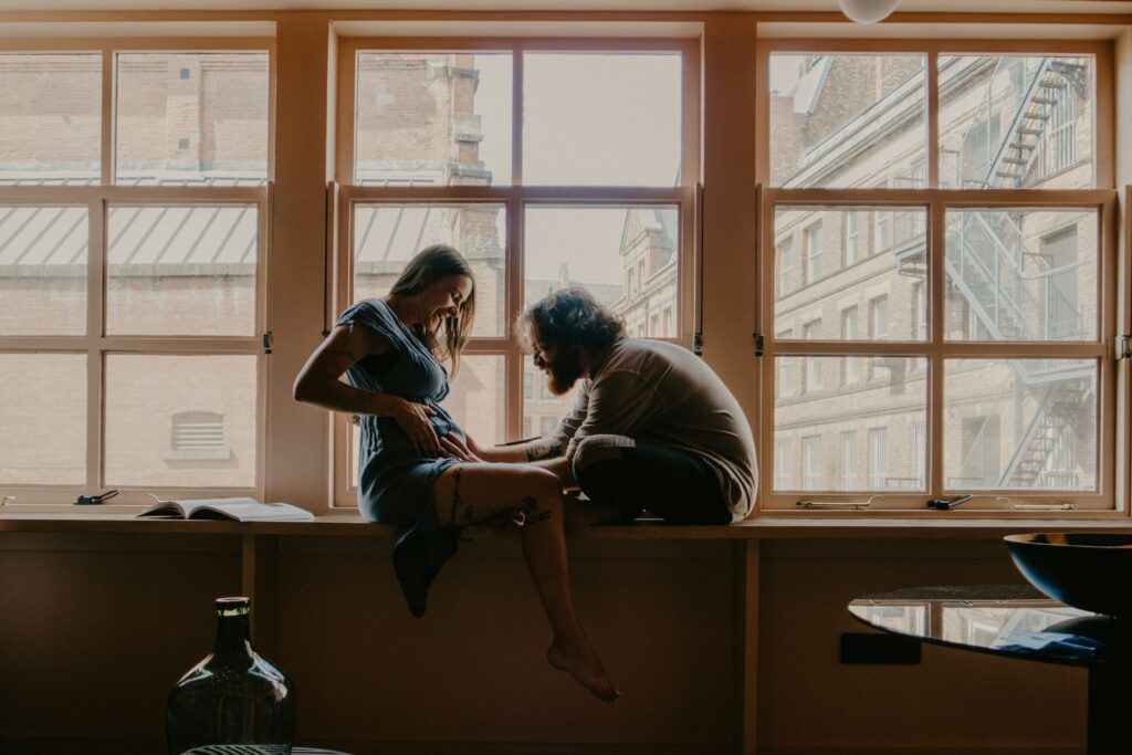 A couple sitting on a windowsill with the man gently touching the woman's pregnant belly as she smiles down at him.