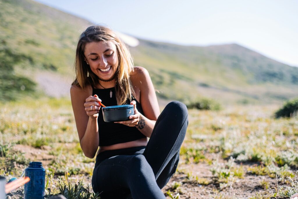 A woman in athletic clothes sits on a grassy hillside next to a bottle of water, smiling while she eats an unknown food from a blue bowl.