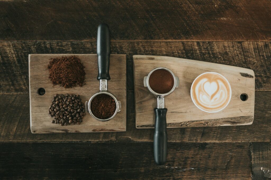 A wooden board on a wooden counter covered in coffee beans, grounds, and espresso tools next to a coffee mug with heart-shaped creamer.