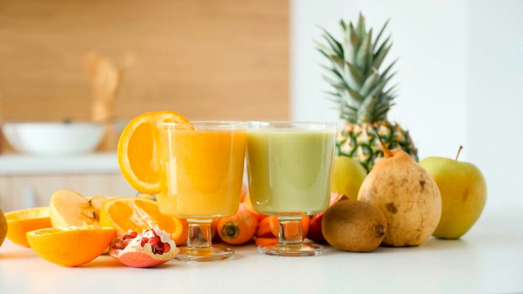 Two glasses of orange and slightly green juice sit on a white counter surrounded by sliced oranges, pomegranates, pears, kiwis and pineapples.