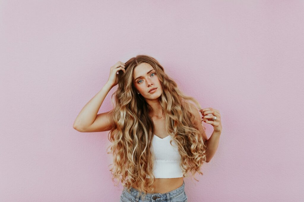 A woman with long curly blonde hair stands in a white crop top and jeans in front of a pink wall. She holds a hand to her hair and her head is tilted to the side.