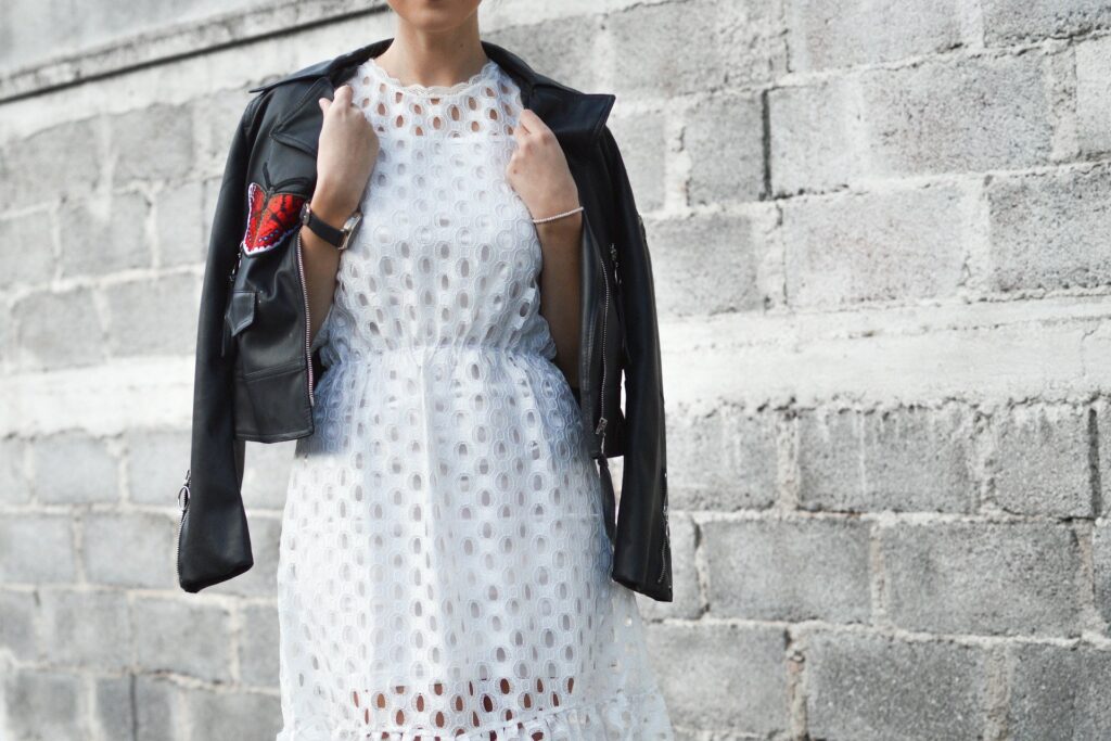 A woman wearing a white dress and a black leather jacket with a red butterfly on it stands in front of a concrete brick wall.