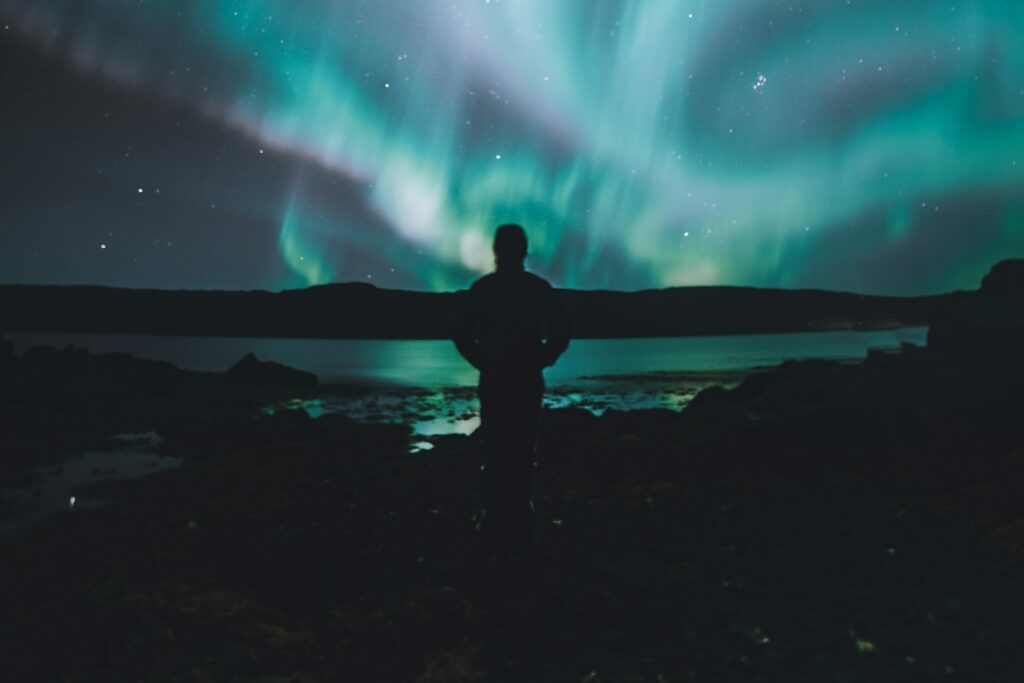 A night owl stands underneath the blue and green Aurora Borealis in the middle of the night. A lake is in front of him and he's on the rocky shore.