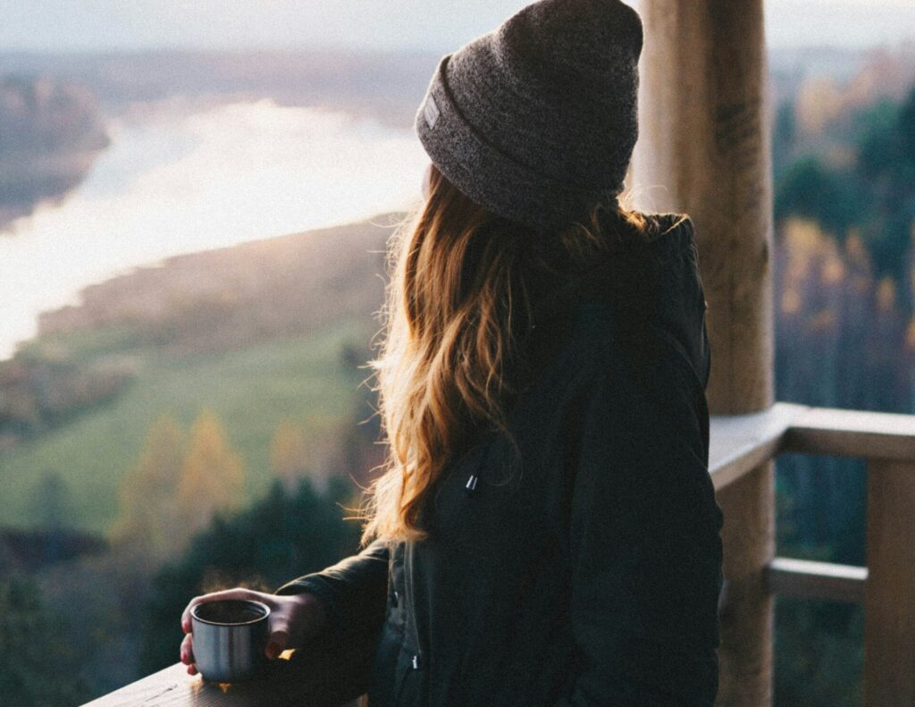 A morning bird stands on a wooden porch overlooking a lake and some mountains at sunrise. She holds a small metal coffee cup that's steaming and she's wearing a knit beanie with a light jacket.