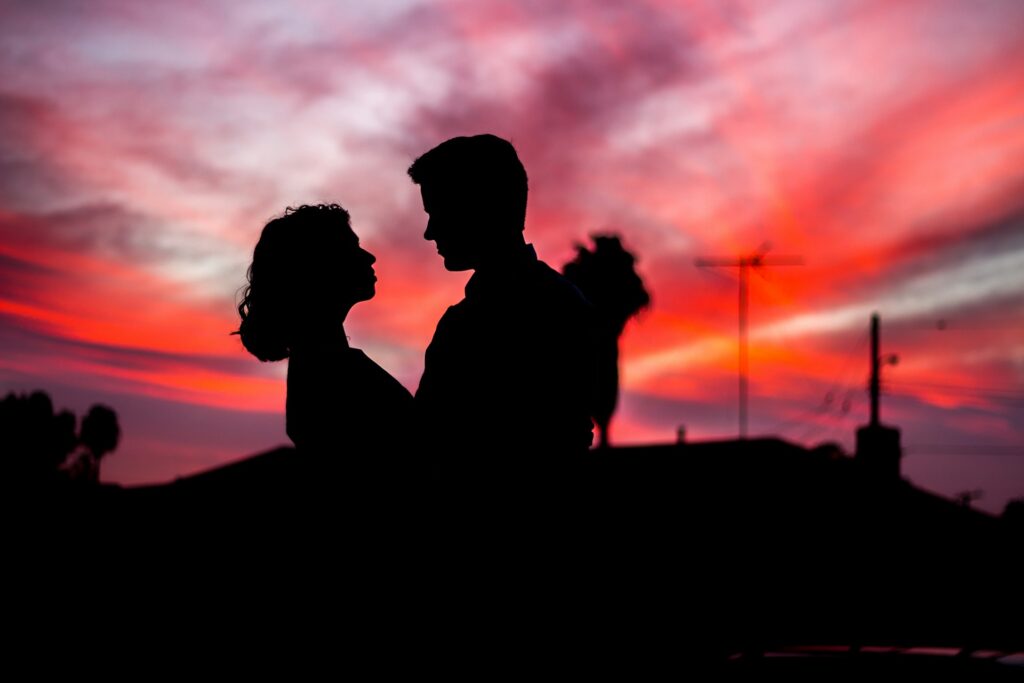 A man and woman embrace in the shadows underneath the pink and purple sky at sunset.