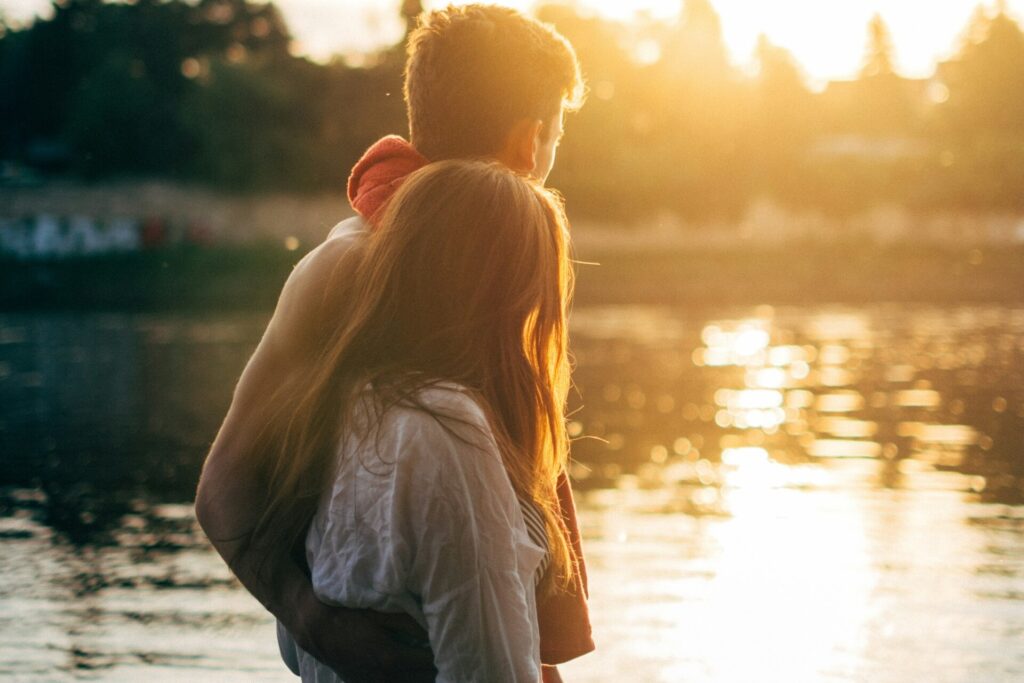 A man and woman hold each other while looking over a lake as the golden sun sets.