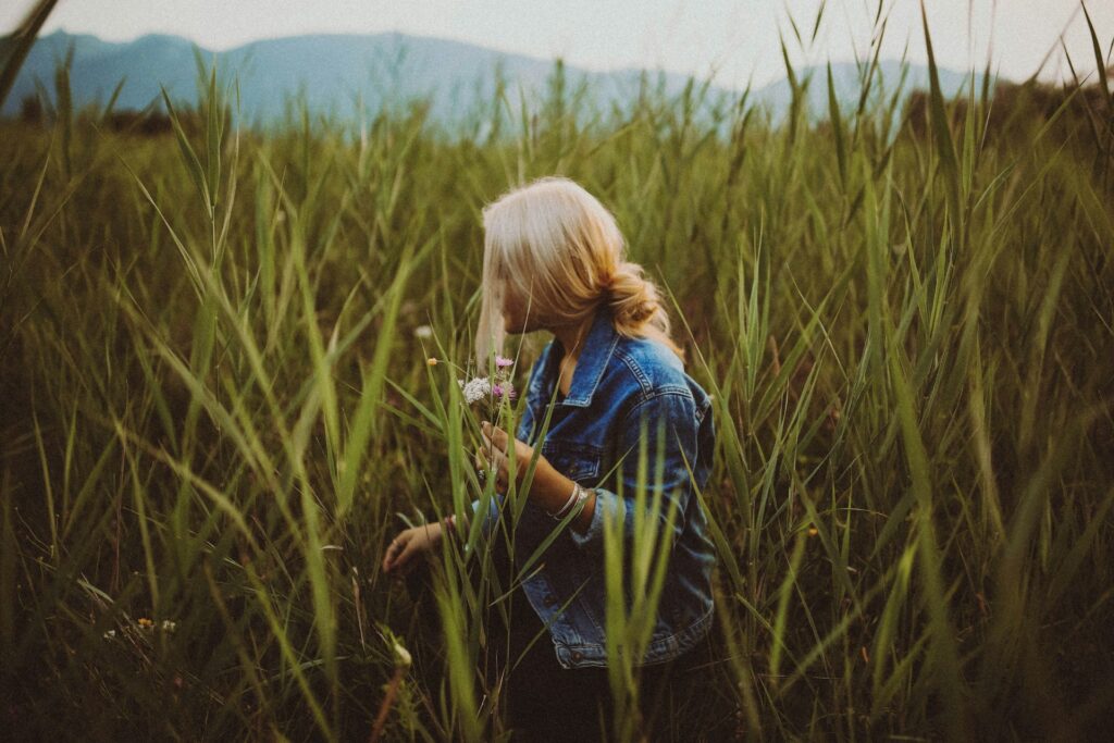 A woman wearing a jean jacket wears her blonde hair in a low messy bun. She holds tiny white and pink flowers in one hand as she navigates a field of green wheat-like stalks that are taller than her. Hills and a gray sky are distant in the background.