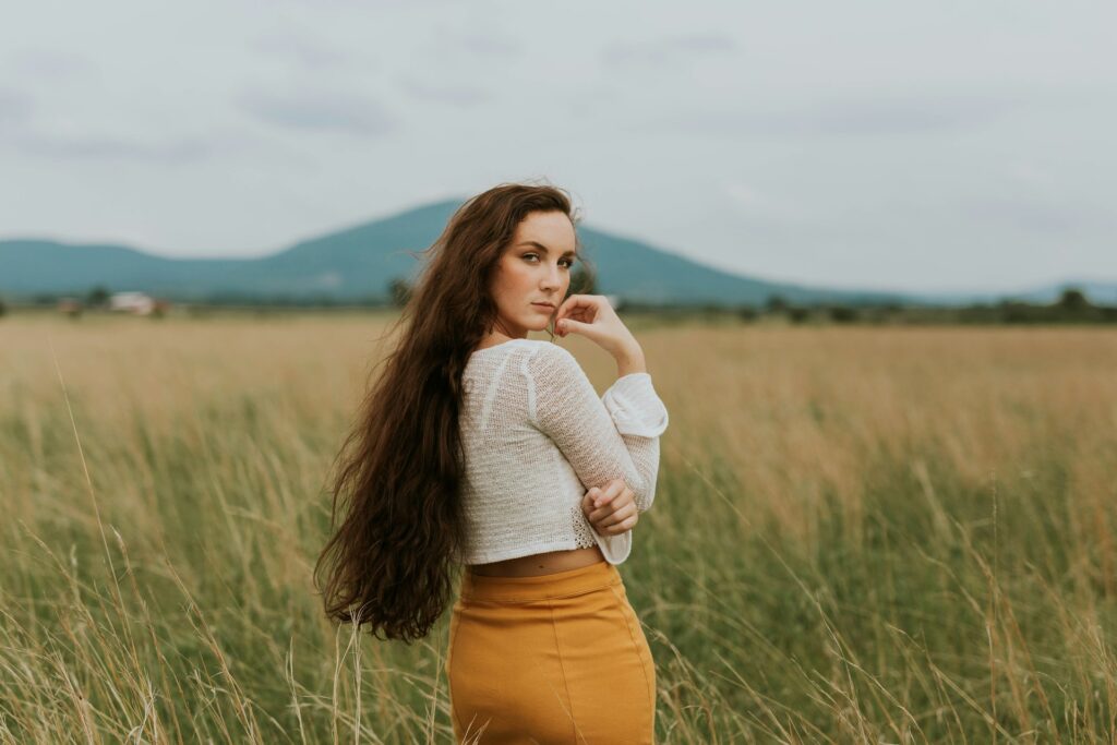 A woman with long brown hair looks back over her right shoulder. She's wearing a white long-sleeved crop top and a yellow pencil skirt. She stands in a wheat field at the base of distant green hills. The sky is full of light gray clouds.