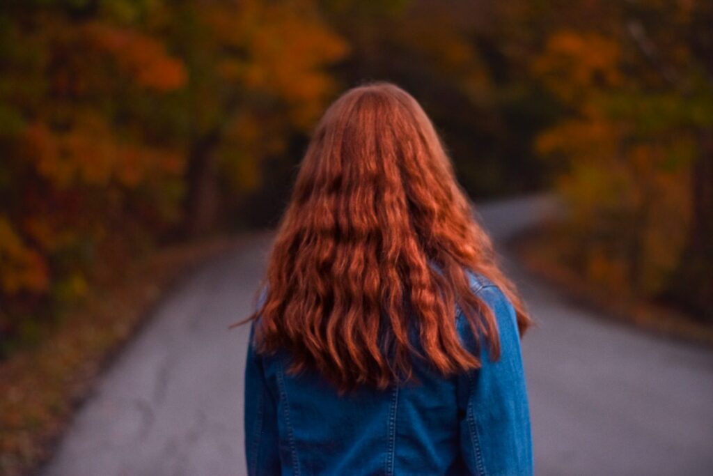 A woman with long red hair and a jean jacket walks on an empty road. The trees on either side are in autumn colors and colorful leaves are scattered on either side of the road.
