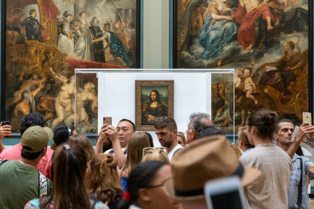 A crowd of people hold up phones in front of the Mona Lisa. Two larger paintings hang on the wall behind it.