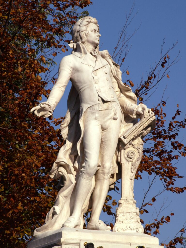 A picture of a marble statue of Mozart in front of a big tree with brown leaves. The sky is blue behind the statue.