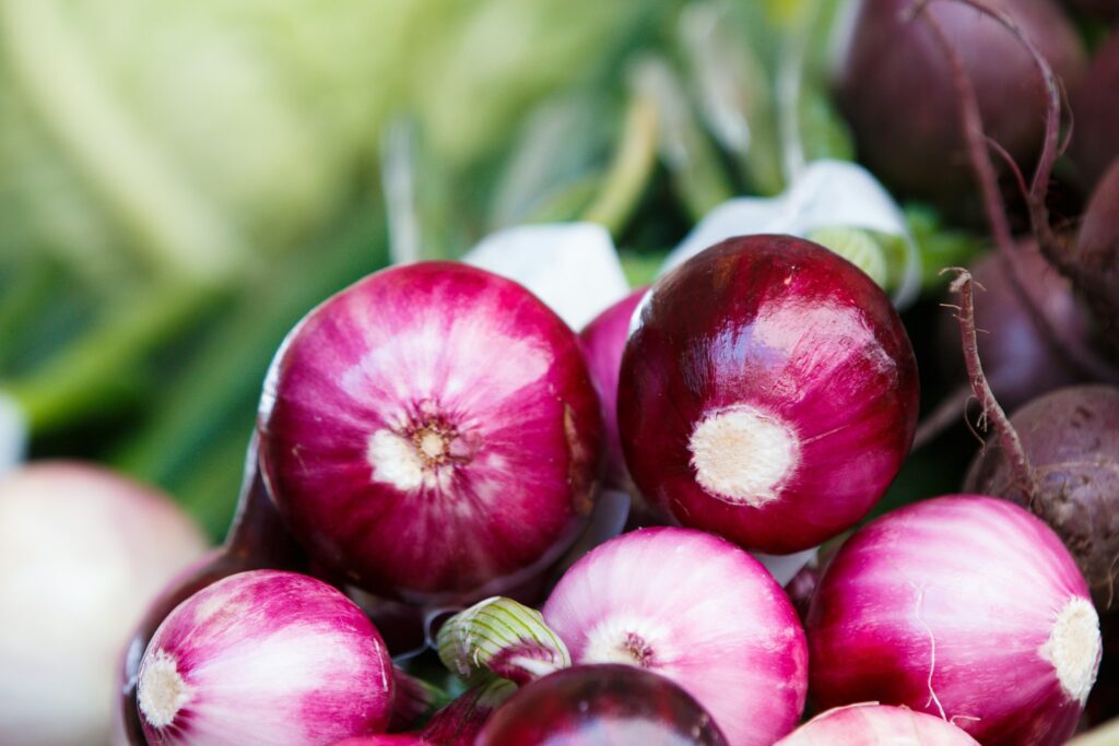 A pile of red onions sits against a background of green vegetables. They're a key ingredient for anyone learning how to make fire cider.