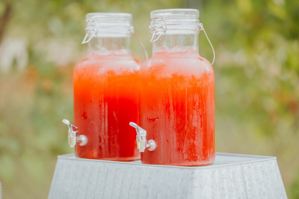 Two glass pitchers with serving spouts sit on an overturned metal bucket against a background of green trees. They contain an orange-red beverage for a party, likely flavored with fire cider.