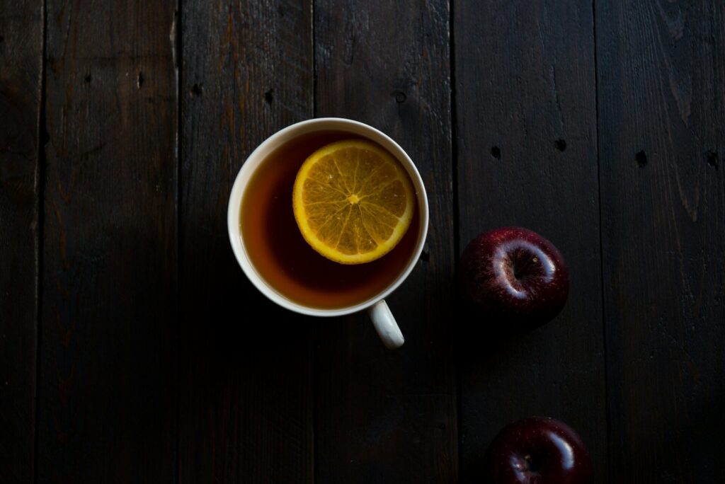 A mug of a brown beverage with a slice of lemon sits on an espresso wood table next to two dark red apples. The beverage is tea flavored with fire cider.