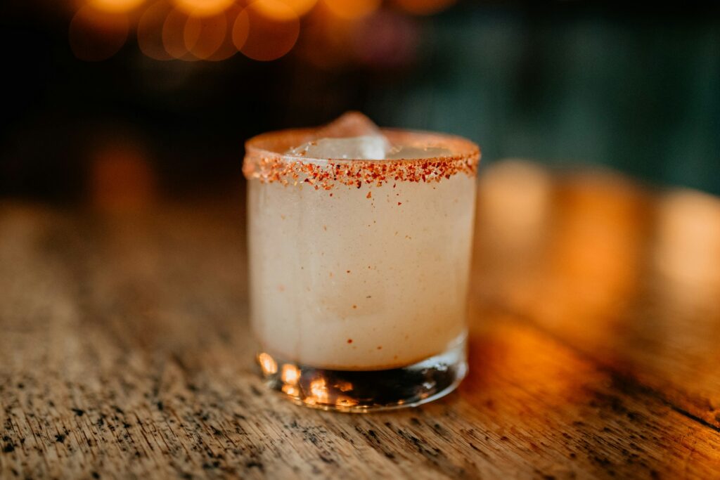 A glass that contains a margarita spiced with fire cider and a cayenne pepper sugar rim sits on a wooden table. Soft orange lights glow in the blurry background.