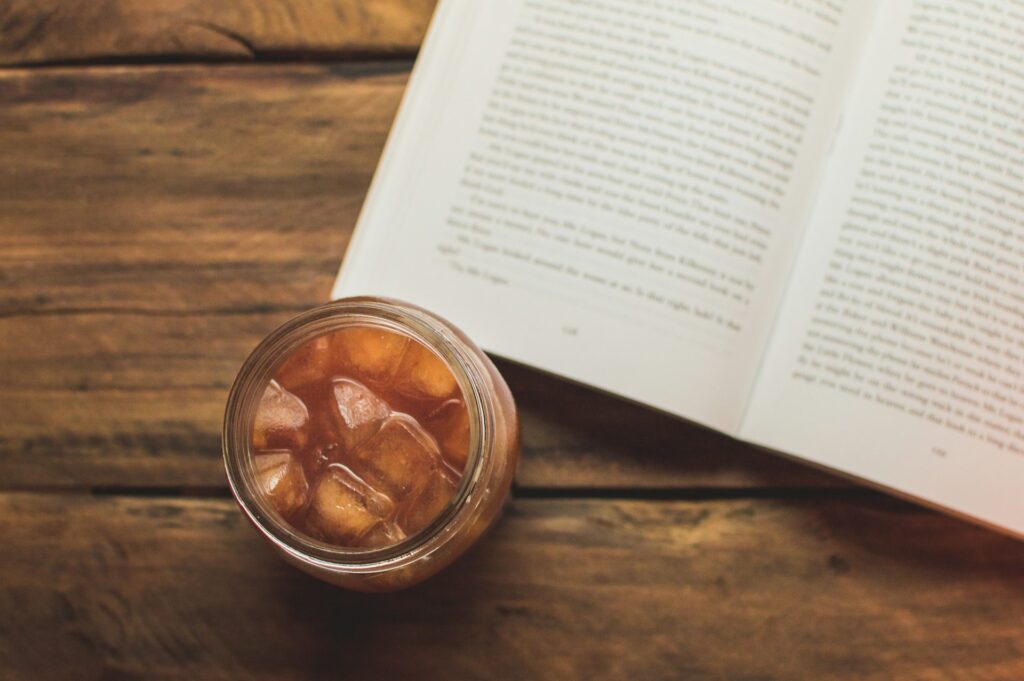 A mason jar of ice cubes and fire cider sits on a wooden table next to an open novel.