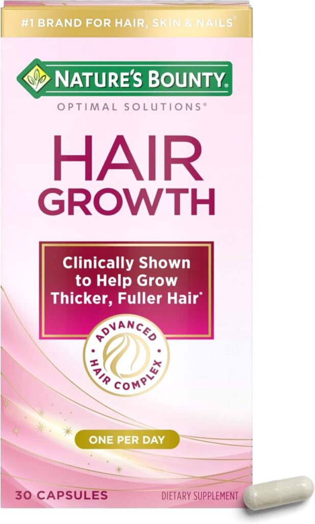 A pink box of Nature's Bounty hair growth supplements sits against a white background. A single white capsule sits in front of it on the right side.