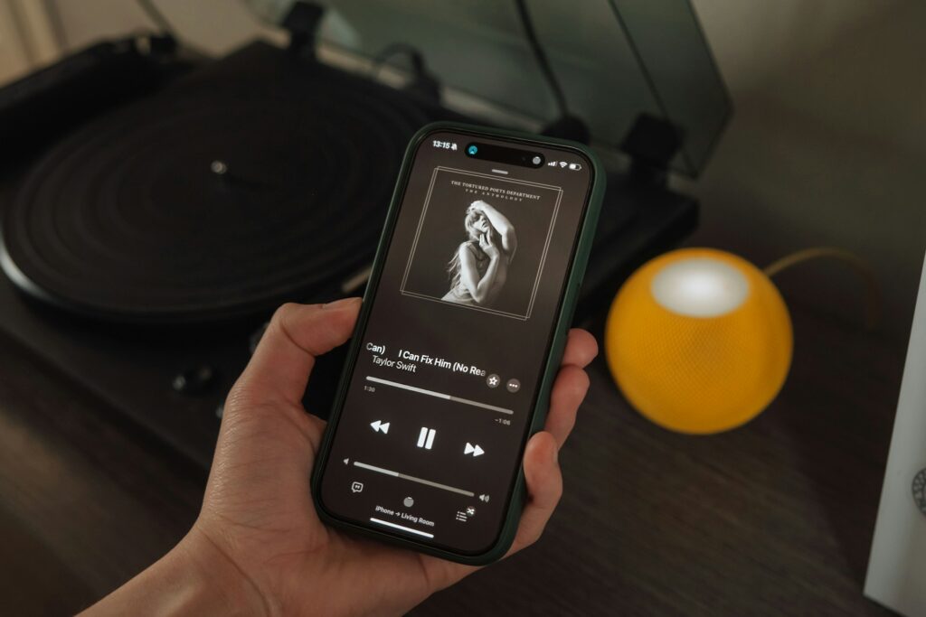 A person holds up their smartphone, which is playing "I Can Fix Him (No Really I Can)" from the The Tortured Poets Department anthology album. The background is an opened, empty record player and a round yellow speaker on a dark wooden dresser.