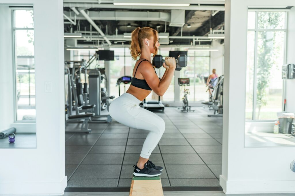 A woman in a black sports bra and white leggings des a crunch on a platform while holding barbells up to her shoulders. The rest of the gym is through a glass doorway behind her. There are multiple types of machinery. One woman uses a standing bike in the distance.