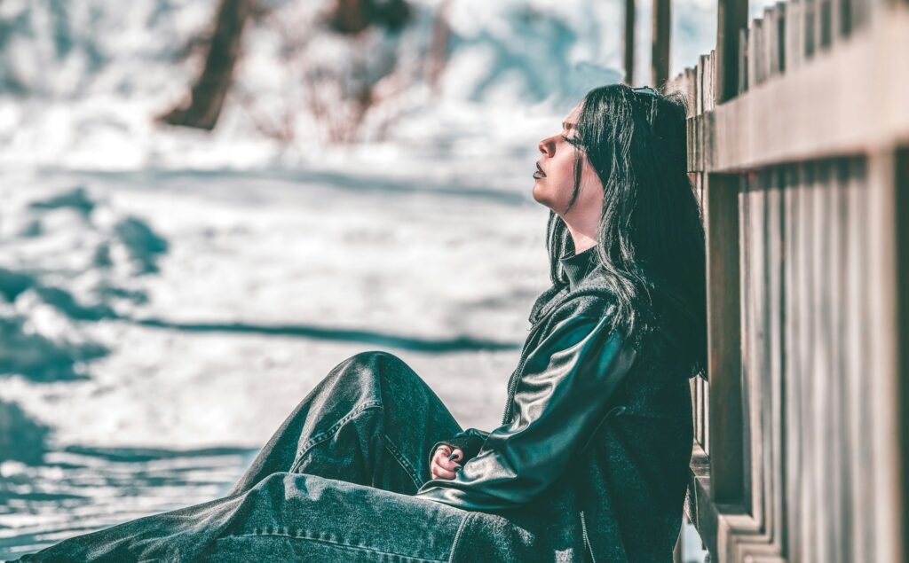 A woman in black jeans and a black leather jacket sits with her back against a fence. Her eyes are closed and her arms are crossed over her midsection like she's uncomfortable.