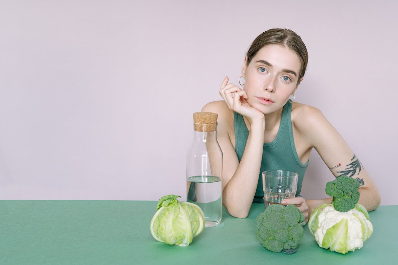 A woman in a green tank top sitting at a green table next to broccoli and cauliflower heads, holding a glass of water