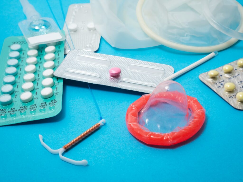 Multiple contraceptive methods sit on a neon blue surface. They include condoms, bills, creams, an IUD and a birth control implant stick.