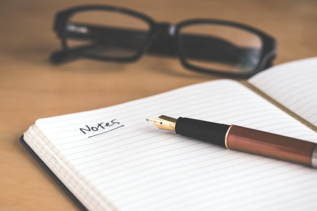 A notebook lies open on a wood surface. The left page says Notes and the word is underlined. A copper fountain pen sits on the page. A pair of black rectangular glasses sits closed on the surface behind the notebook. It could be used while someone learns what happens when you get off birth control.