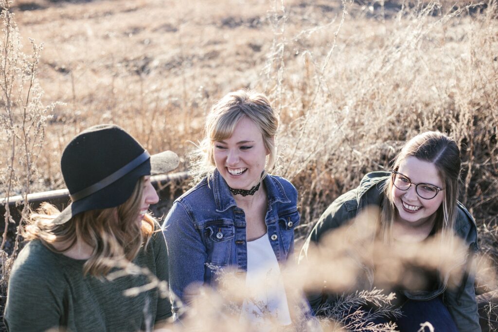 Three women sit on a bench in front of tall brown grasses. It's sunny and they're all laughing together.