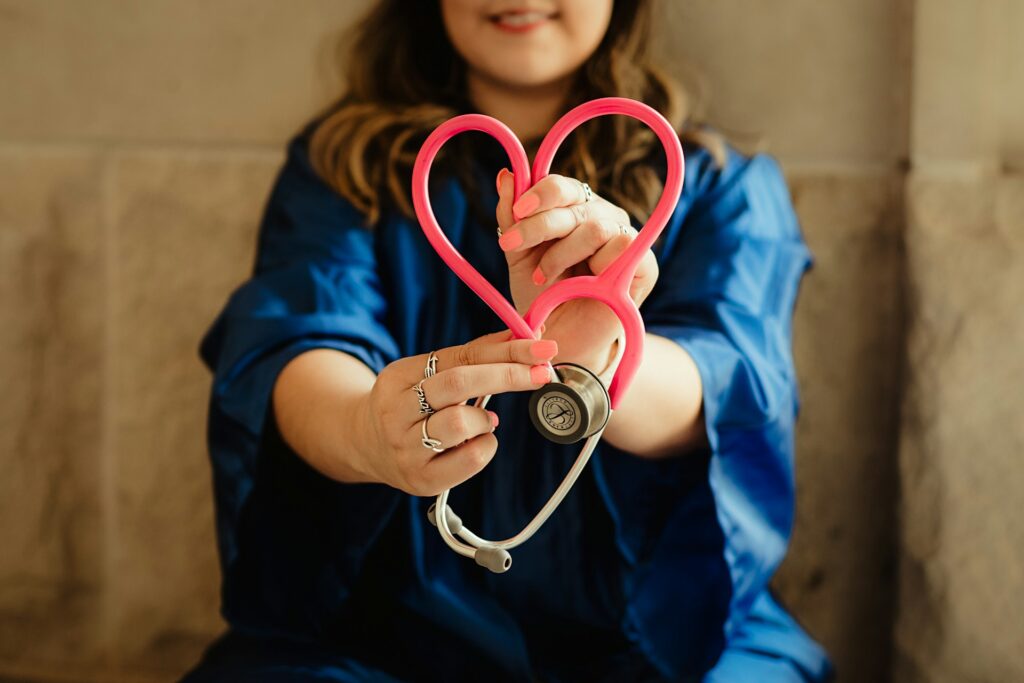 A woman holds a pink stethoscope bent in the shape of a heart toward the camera.