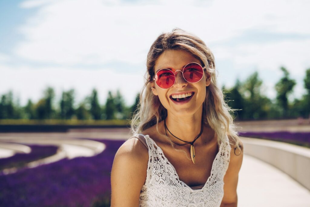 A blonde woman in a white lace tank top and red circular sunglasses sits on a small concrete wall among purple flowers. She's smiling and laughing.