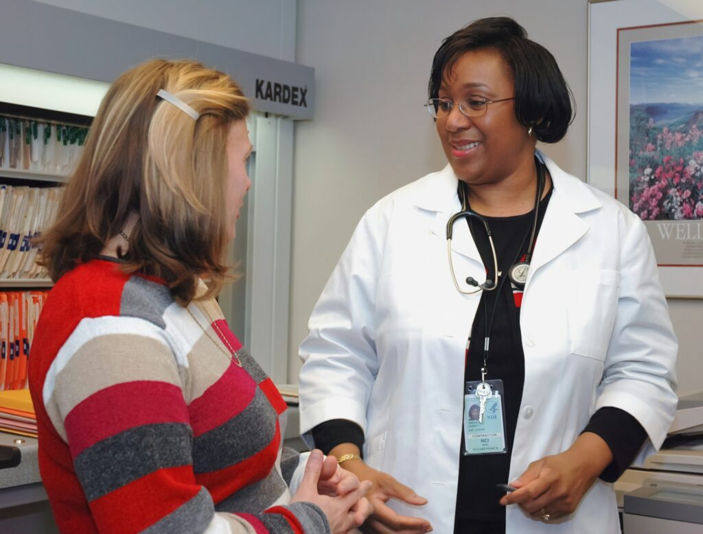 A woman in a red striped shirt talks with a doctor wearing a white coat. A wall of patient files is behind them.