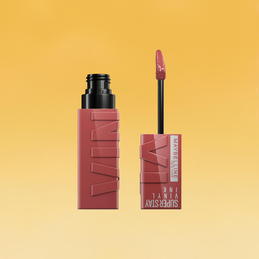A pink tube of Maybelline's Super Stay Vinyl Ink liquid lipstick sits against the yellow Body+Mind background. The tube is on the left and the handle brush is on the right. It's among the best drugstore beauty products.