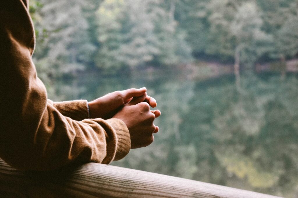 A person wearing an orange sweater leans on a wooden railing overlooking a lake. The water reflects the many green trees surrounding the lake.