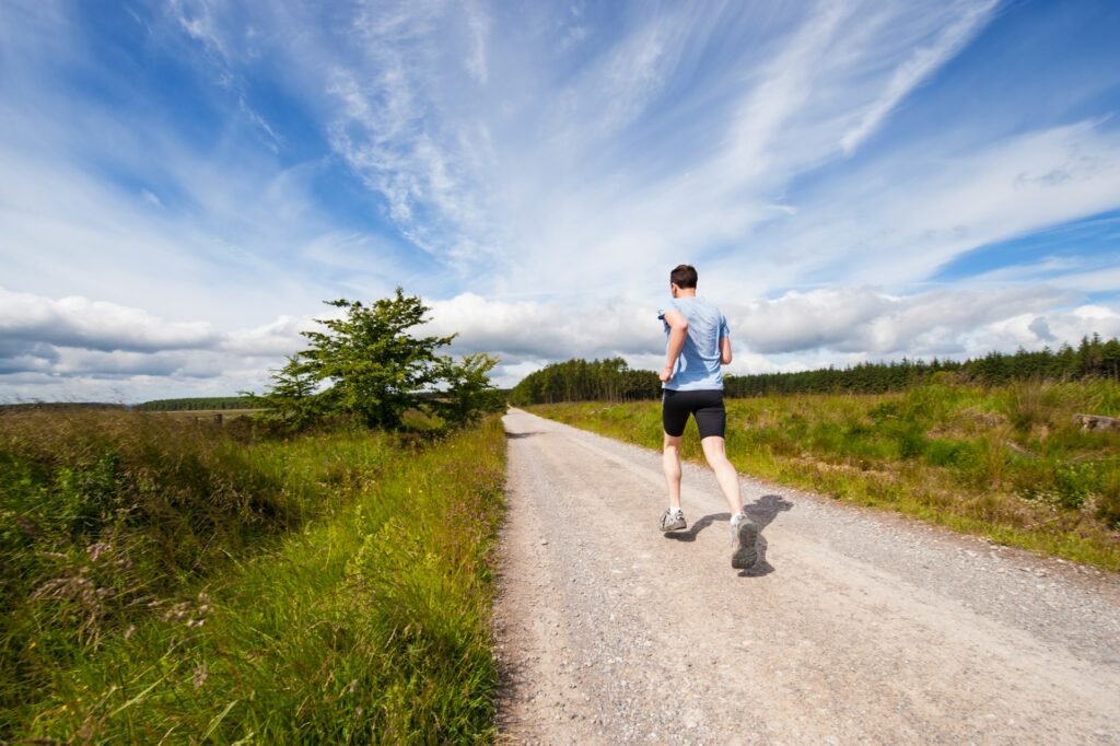 A man in black shorts, a blue shirt and white shoes runs down a gravel road under a blue sky. Empty green fields and forests stretch out on either side of him.