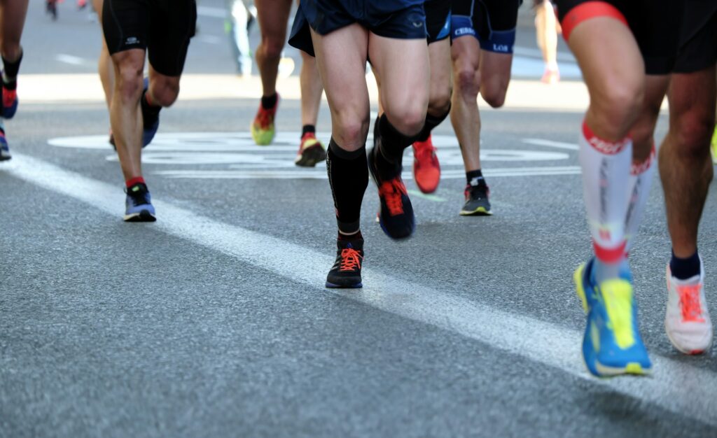Seven runners race across a road. Only their tennis shoes and legs are visible. Everyone is wearing multi-color clothes and shoes.