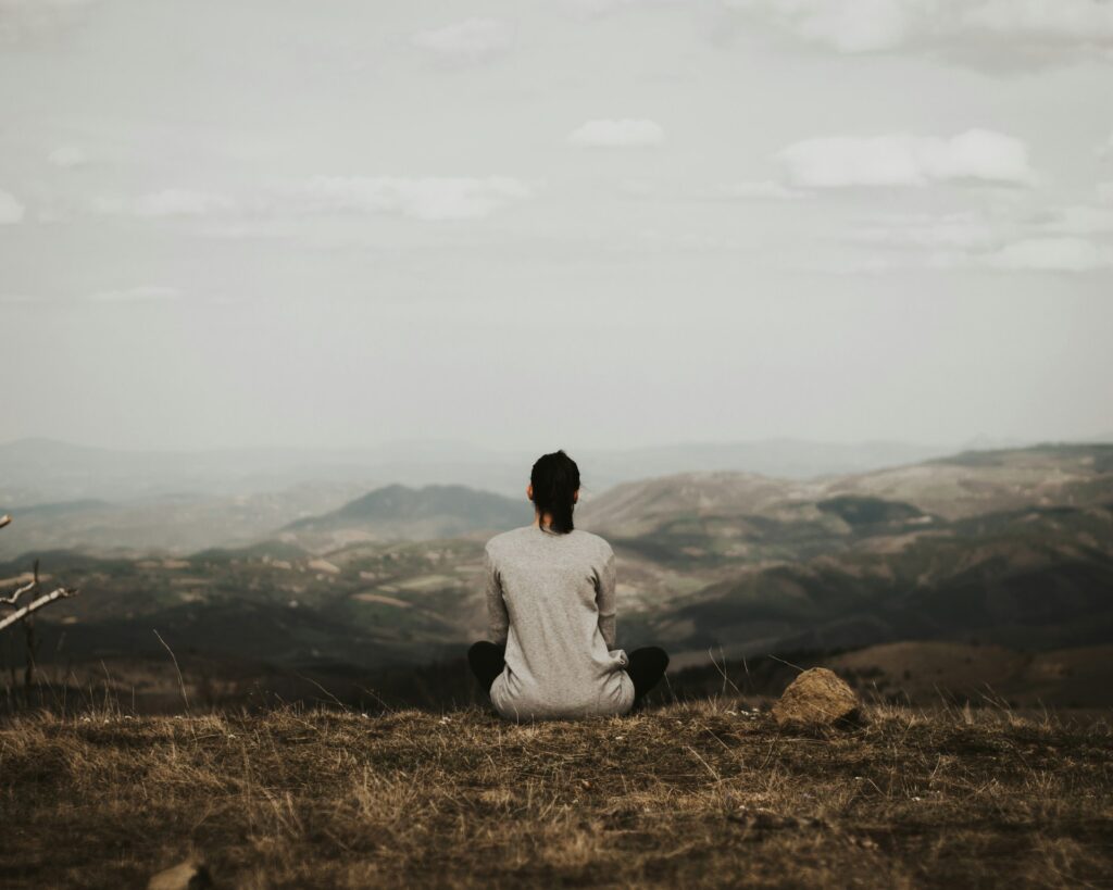 A woman sits cross legged on top of a hill overlooking fields, hills and a small village. The sky is gray and overcast.