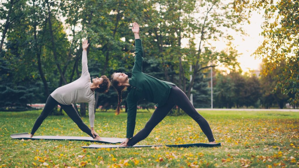 Two women bend over doing yoga on black mats in a public park. Yellow leaves are across the ground and the sun is setting behind them. They might be doing a workout for PCOS.