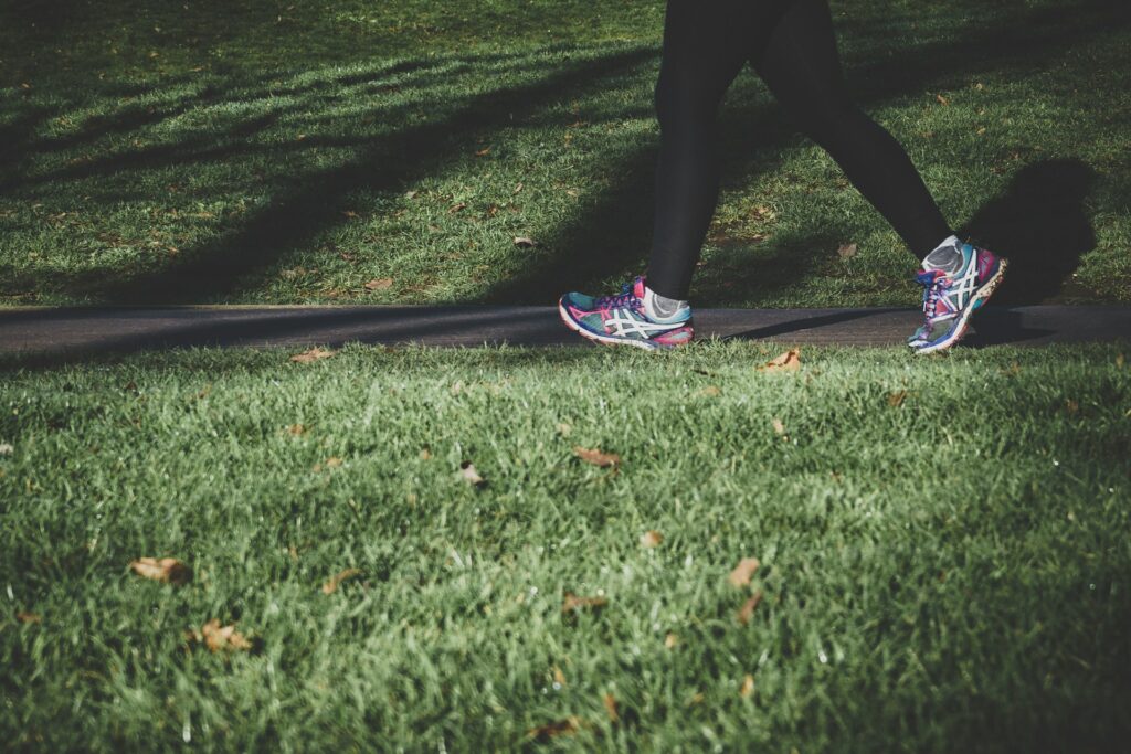 A woman in black leggings and multi-colored tennis shoes jogs down a concrete sidewalk among green grass.