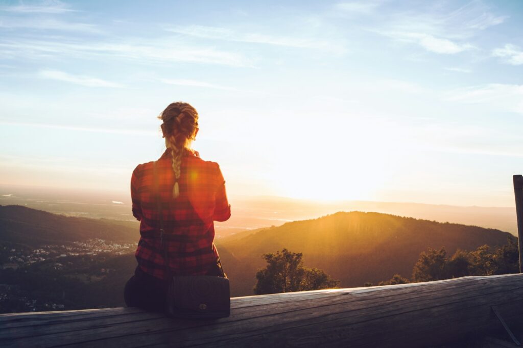 A woman with fishtail braided hair sits on a wooden bench overlooking a serene mountain sunset. She's wearing a red plaid shirt, conveying peace and reflection.