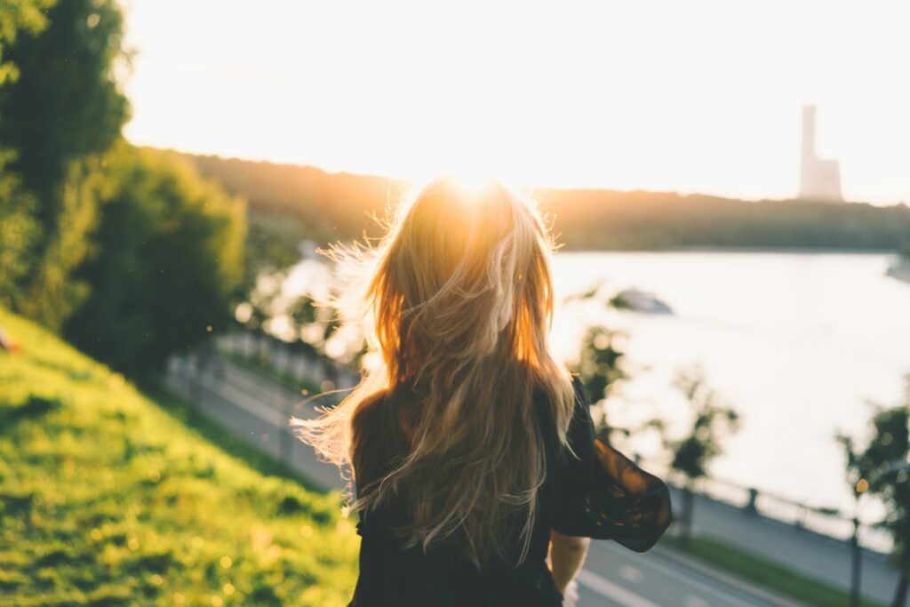 A blonde woman in a black shirt walks across a grassy hillside. The sun is setting. Two roadways are at the bottom of the hill, next to a lake with a tall factory beyond.