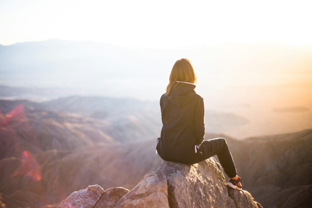 A woman in black pants and a black windbreaker sits on a rocky hilltop overlooking a valley. The sun sets in front of her.