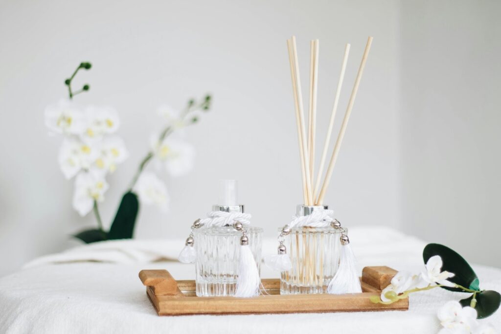 A wooden tray holds a glass reed diffuser with sticks and a matching glass spray bottle with white tassels, white orchids are in the background.